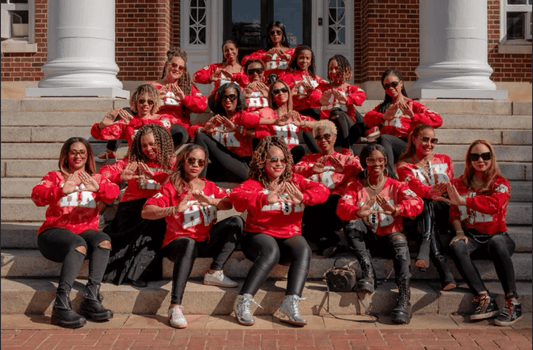 Group of Delta Sigma Theta Sorority members sitting together in front of their sorority house, wearing matching Delta Sigma Theta shirts and holding up a sorority sign.