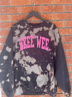 Skee-Wee sweatshirt with pink text on a hanger against a brick wall.