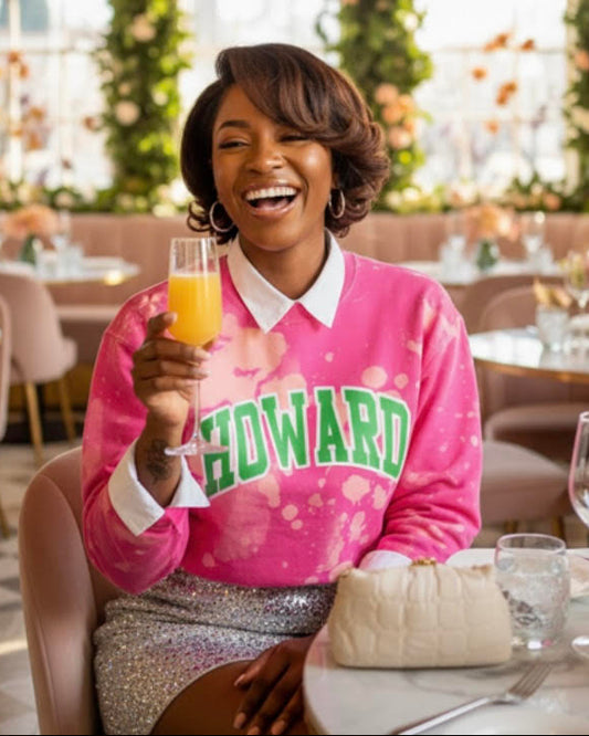 A stylish young Black woman wearing a hand-bleached pink sweatshirt with “HOWARD” in bold green lettering, smiling and holding a mimosa while seated in a floral, pastel-toned brunch restaurant.