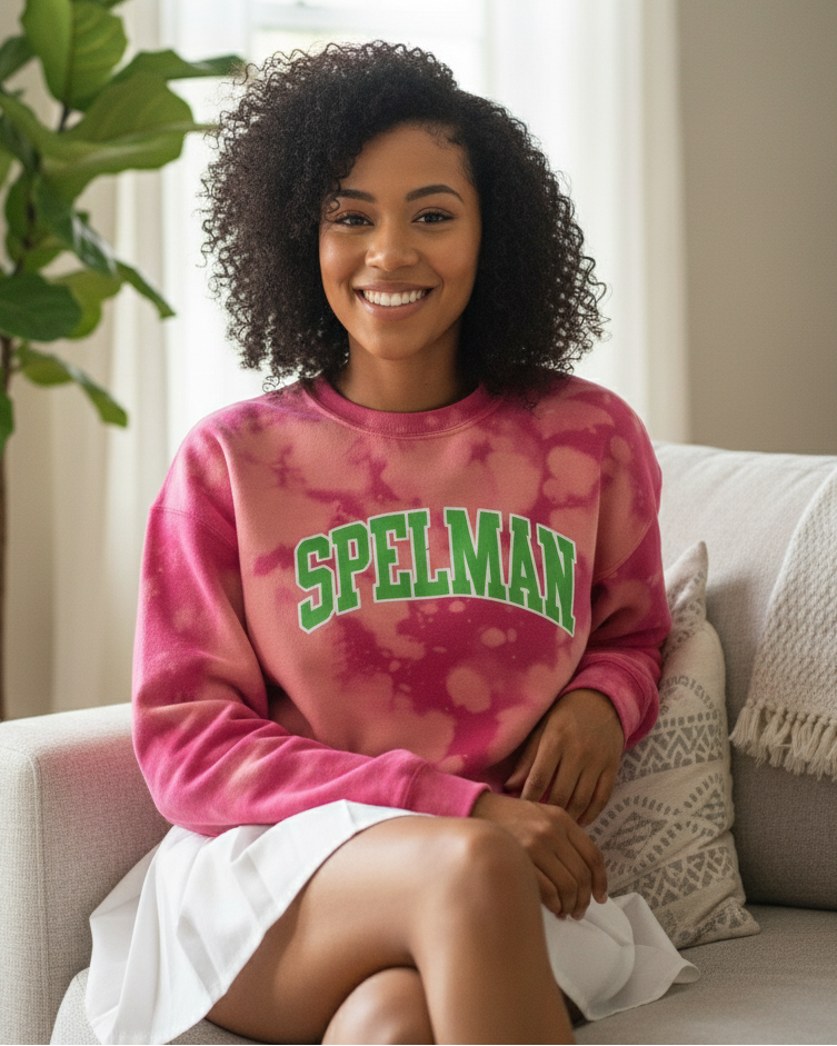 A smiling young Black woman with natural curly hair sitting cross-legged on a white sofa wearing a pink hand-bleached sweatshirt with the word “LEGACY” in bold green arch lettering, styled with a white pleated skirt in a cozy, sunlit living room.