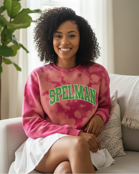 A smiling young Black woman with natural curly hair sitting cross-legged on a white sofa wearing a pink hand-bleached sweatshirt with the word “LEGACY” in bold green arch lettering, styled with a white pleated skirt in a cozy, sunlit living room.